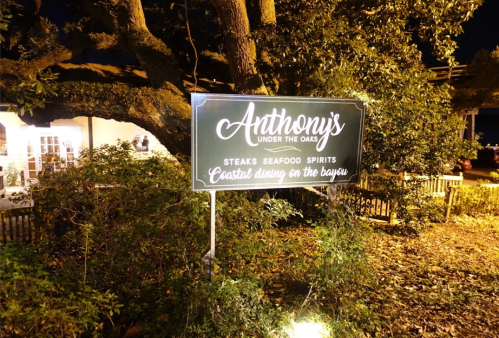 Sign for "Anthony's Under the Oaks" restaurant, featuring steaks, seafood, and spirits, set in a bayou location at night.