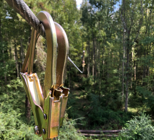 Close-up of a metal zip line harness on a cable, with a lush green forest in the background.