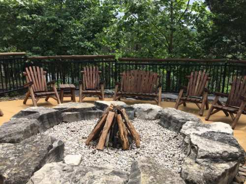 A circular stone fire pit surrounded by wooden chairs on a deck, with lush greenery in the background.