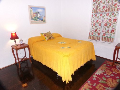 Cozy bedroom with a yellow bedspread, floral curtains, a bedside lamp, and a small table.