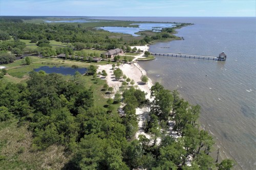 Aerial view of a coastal landscape with a sandy shore, green trees, a pond, and a pier extending into the water.