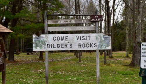 Sign welcoming visitors to Bilgers Rocks, featuring a message to come visit and a no alcohol notice. Surrounded by trees.