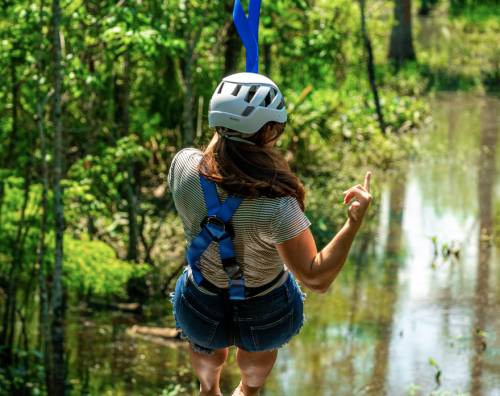 A person in a helmet zip-lining over a lush, green landscape with a reflective water surface below.