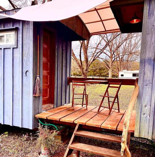 A cozy blue tiny house with a wooden porch and two chairs, surrounded by trees and a grassy area.