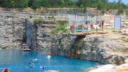 A sunny swimming area with people diving and swimming in a blue quarry lake surrounded by rocky cliffs.