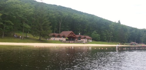 A serene lakeside view featuring a wooden lodge surrounded by trees and a sandy beach.