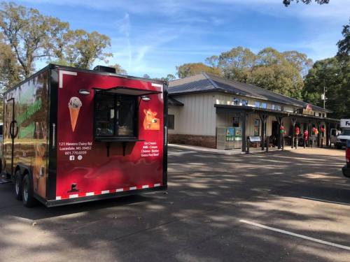 A red food truck parked outside a building, with people lined up in front, surrounded by trees.