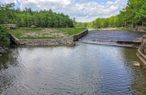 A serene landscape featuring a calm river, a small waterfall, and lush green trees under a partly cloudy sky.