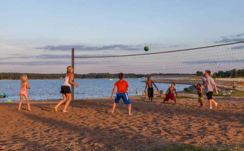 Children playing beach volleyball near a lake during sunset, with a sandy shore and water in the background.