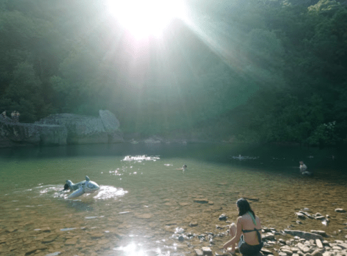 A sunny day at a natural swimming area with people swimming and relaxing by the water, surrounded by greenery.