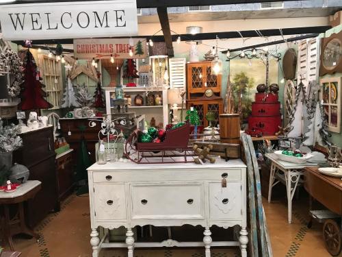 A cozy shop display featuring vintage decor, Christmas trees, and festive ornaments, with a welcoming sign above.