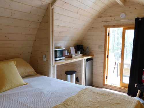 Cozy wooden cabin interior with a bed, kitchenette, and large windows opening to a forest view.