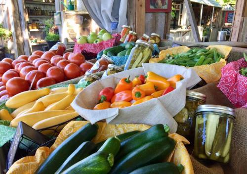 A vibrant display of fresh vegetables and jars of pickles at a market, featuring tomatoes, squash, peppers, and zucchini.