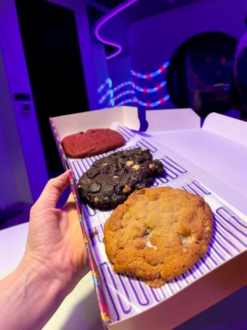 A hand holding a box of three large cookies: red velvet, chocolate chip, and a dark cookie, against a colorful background.