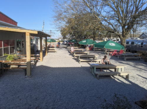 Outdoor seating area with picnic tables and green umbrellas, surrounded by gravel and trees under a clear blue sky.