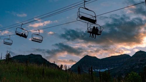 Silhouettes of people on a ski lift against a sunset sky and mountain backdrop.
