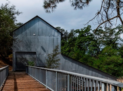 A weathered gray barn with a sloped roof, surrounded by trees, viewed from a wooden walkway.