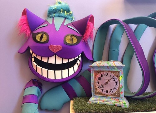 A colorful, oversized cat face with a big grin and a whimsical clock beside it, set against a purple background.