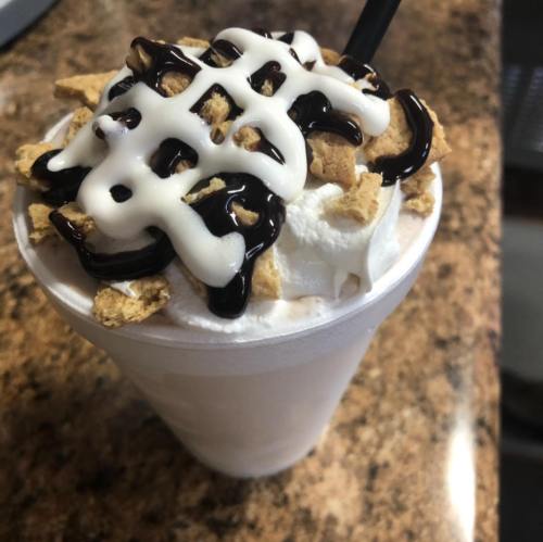 A creamy dessert drink topped with whipped cream, chocolate drizzle, and crunchy cereal pieces in a cup.