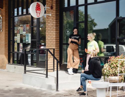 Two women chat outside an ice cream shop, one sitting on steps and the other standing, both enjoying drinks.