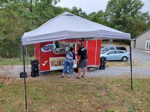 A food truck under a canopy with an "OPEN" sign, two people ordering, and a car parked nearby in a wooded area.