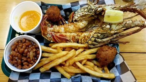 A plate featuring grilled lobster, fries, baked beans, and two hushpuppies, garnished with a lemon wedge.
