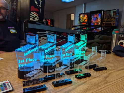 A row of illuminated trophies labeled 1st, 2nd, and 3rd place, displayed on a table with pinball machines in the background.