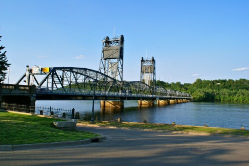 A metal drawbridge spans a river, with green trees and a clear blue sky in the background.