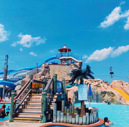 A water park scene featuring a large slide, a lifeguard tower, and people enjoying the pool under a blue sky.
