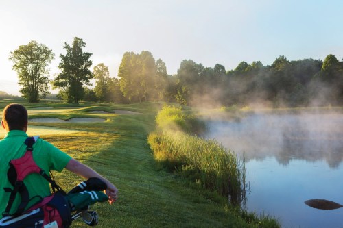 A golfer walks along a misty lakeside path at dawn, with lush greenery and a serene golf course in the background.