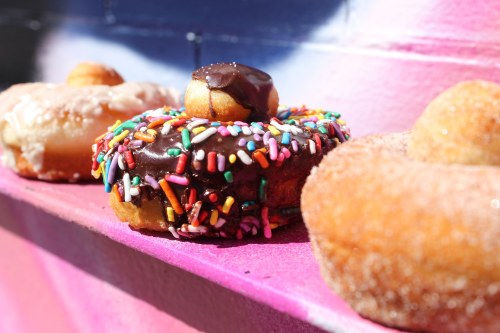 A colorful donut topped with chocolate and sprinkles, alongside two other glazed donuts on a vibrant pink surface.