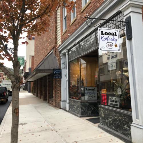 A storefront with a "Local Kentucky" sign, featuring a tree and brick buildings along a city sidewalk.