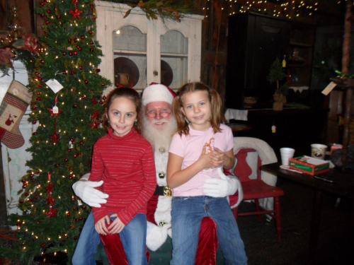 Two girls sit on Santa's lap in a festive room decorated for Christmas, with a tree and holiday decorations in the background.