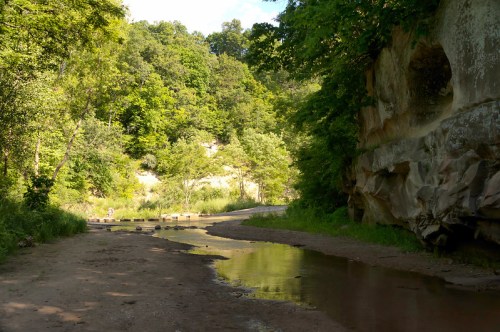 A serene landscape featuring a sandy path beside a calm stream, surrounded by lush green trees and rocky cliffs.