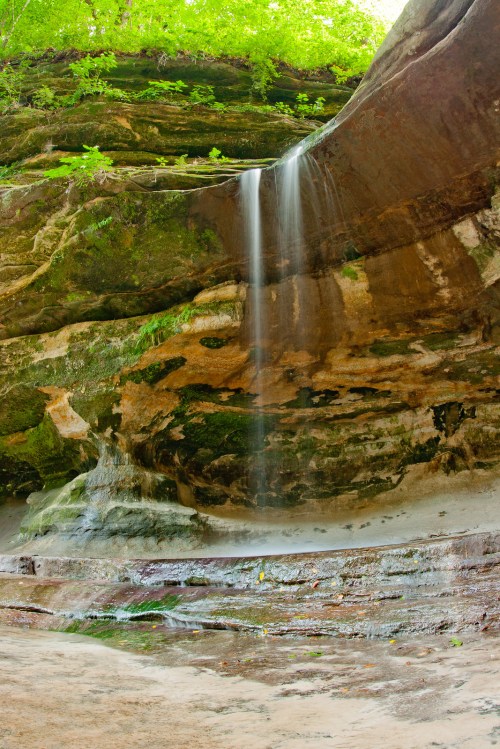 A serene waterfall cascades over moss-covered rocks in a lush, green forest setting.