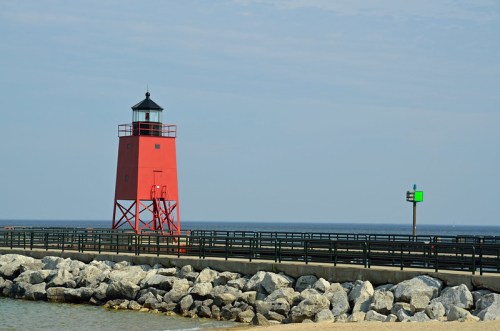 A red lighthouse stands on a rocky pier, with a green navigation light in the background and calm waters beyond.