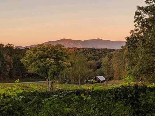 A serene landscape featuring rolling hills, a rustic barn, and trees with autumn foliage under a soft sunset sky.