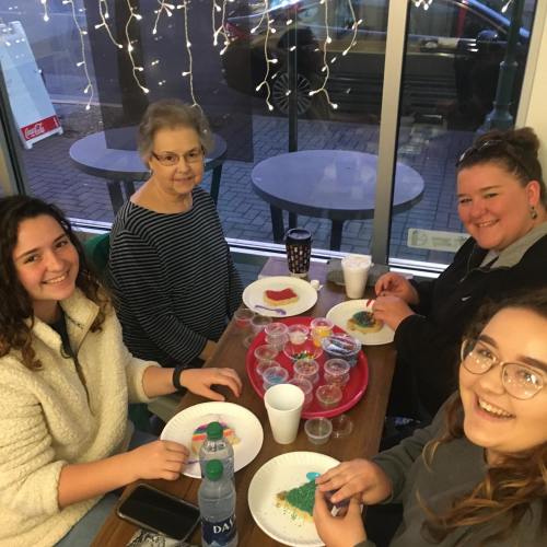 Four people sitting at a table, decorating cookies with colorful toppings, surrounded by festive lights.