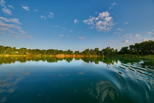 A serene lake reflecting a blue sky with scattered clouds and lush greenery along the shore.