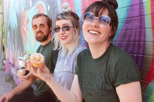 Three friends sit against a colorful mural, smiling and enjoying donuts while wearing matching green shirts.
