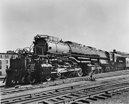A vintage steam locomotive parked on tracks, with a clear sky and buildings in the background.