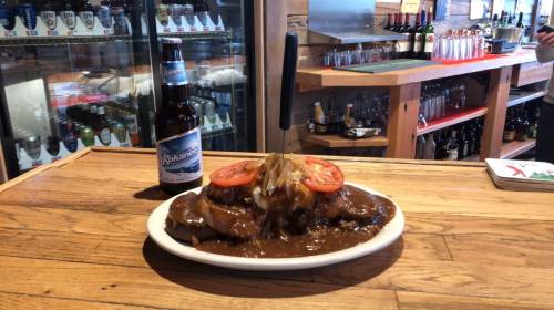 A plate of meat topped with tomatoes and onions, served with a beer, on a wooden bar counter.