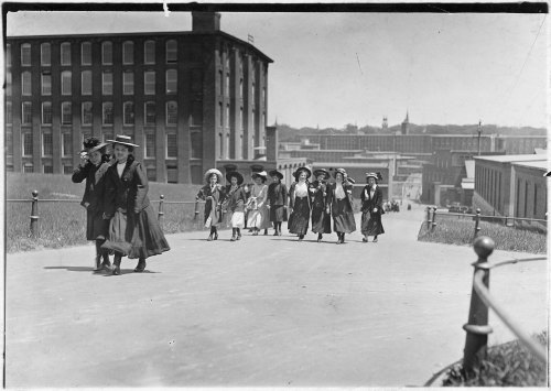 A group of women in early 1900s attire walking down a road, with buildings and a cityscape in the background.
