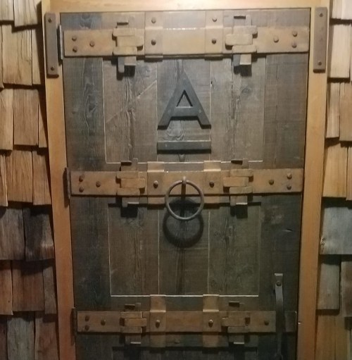 A rustic wooden door with metal hardware and an "A" symbol, set against a textured wooden wall.