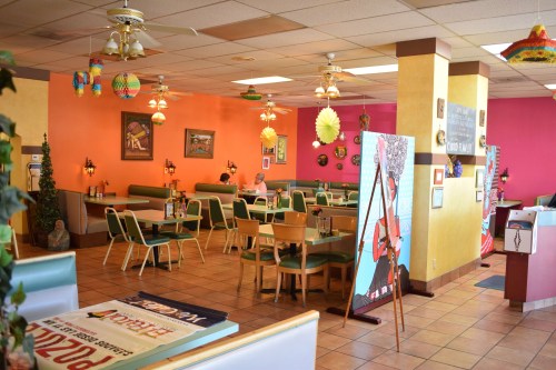 A colorful restaurant interior with empty tables, vibrant decor, and a person sitting alone at a table.