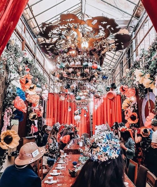 A vibrant indoor space decorated with large flowers, hanging art, and colorful hats, filled with people at a long table.