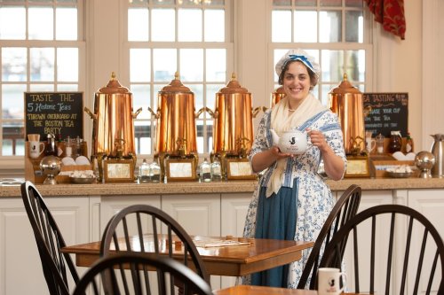 A woman in historical attire holds a teapot in a tea room with copper tea kettles and chalkboard menus in the background.