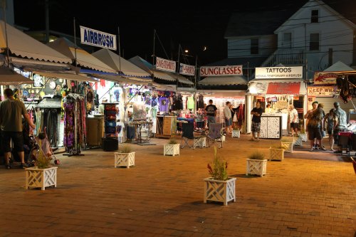 Night market scene with various vendor tents selling airbrush art, sunglasses, and tattoos, surrounded by shoppers.