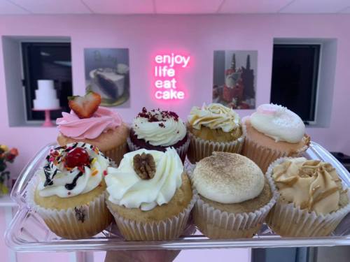 A tray of colorful cupcakes with various toppings, displayed in a pink-themed bakery with a neon sign.