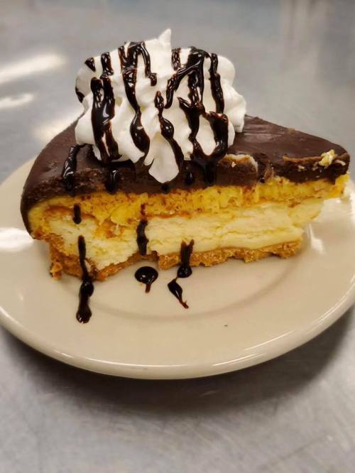 A slice of layered dessert with a chocolate topping, whipped cream, and chocolate drizzle on a plate.